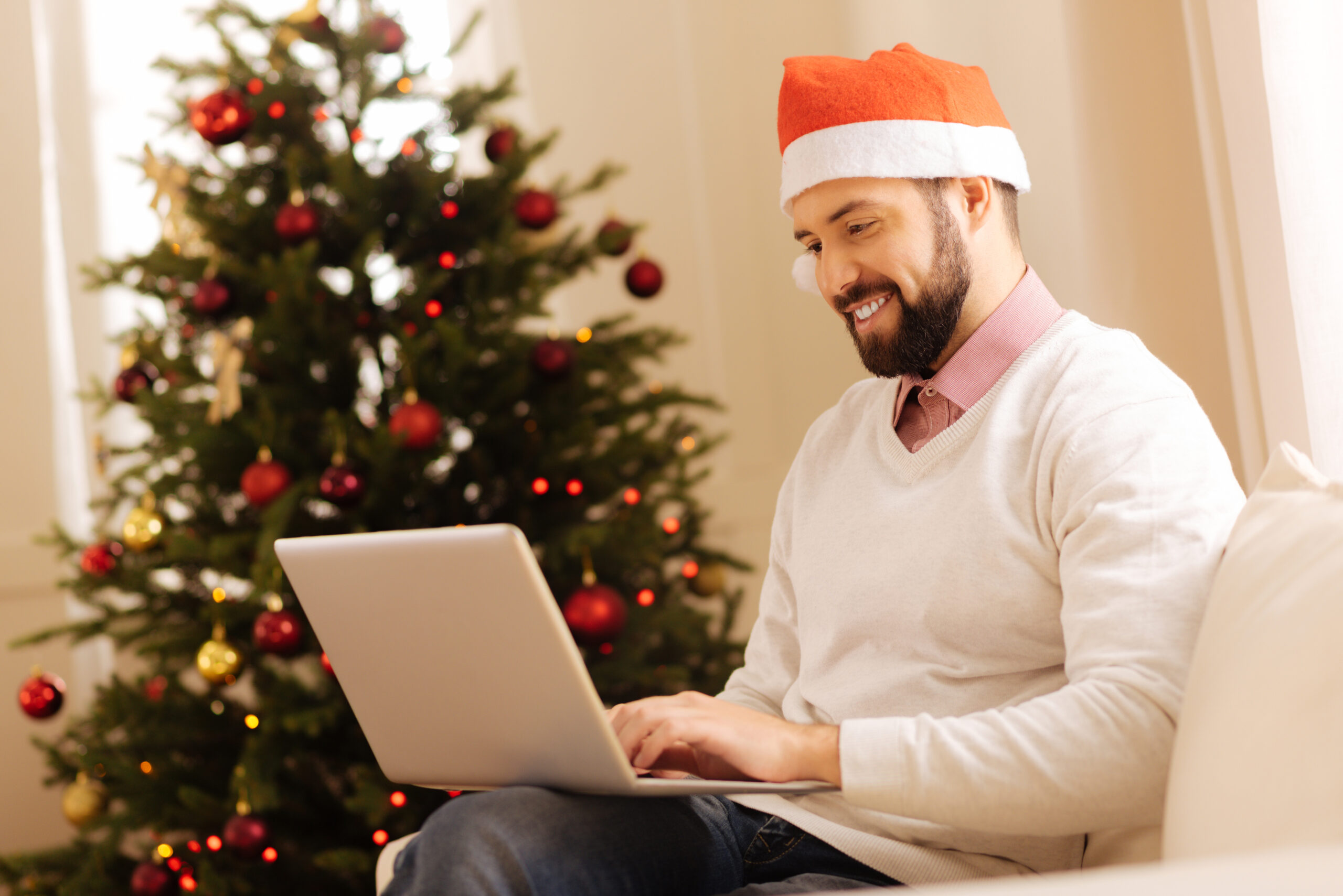 Christmas mood. Charming young man sitting on the sofa in the room with Christmas tree and working on the laptop while wearing a Santa hat.
