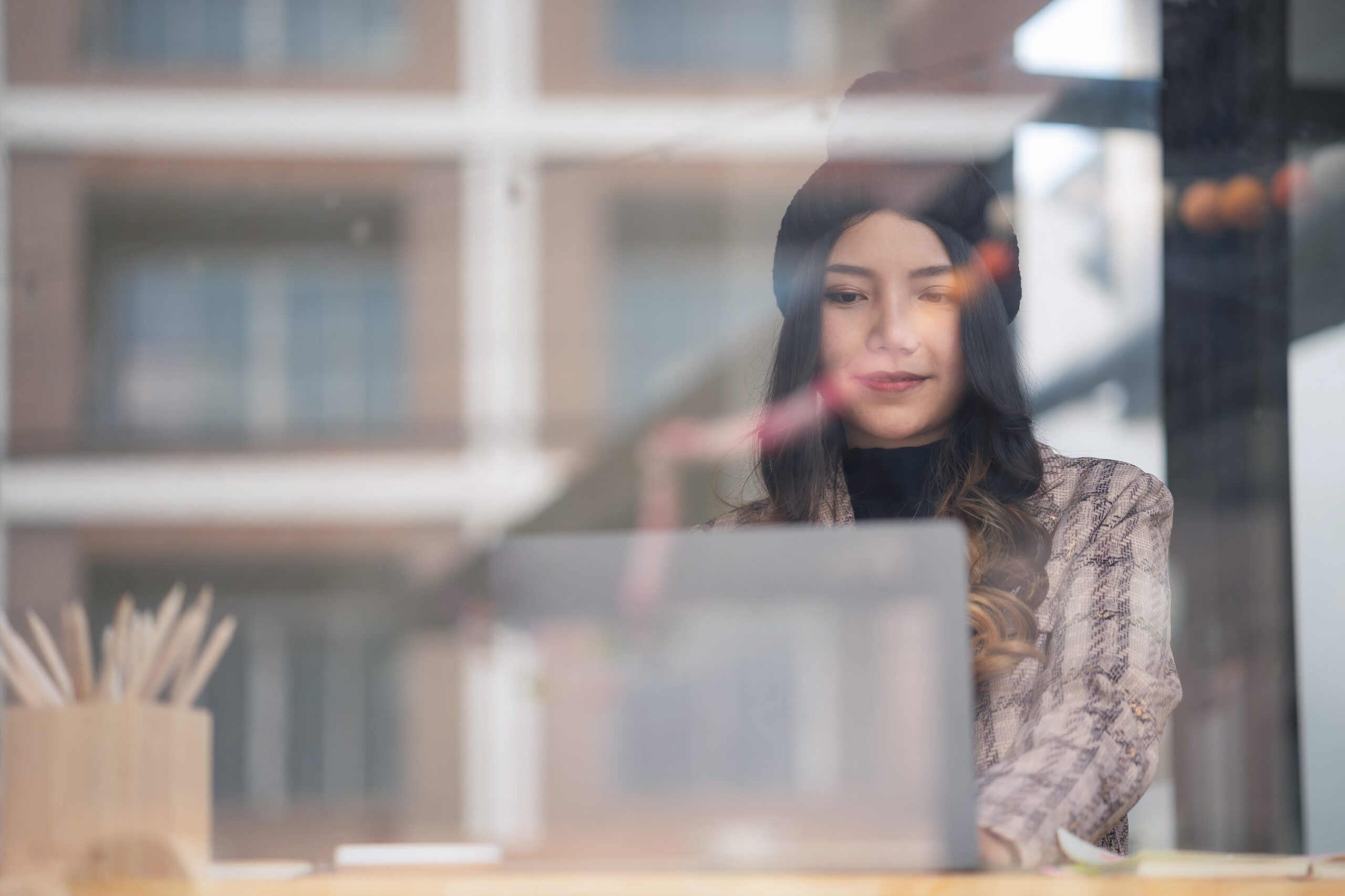 Glass view of female college student wearing winter knitwear sitting and working with a laptop.