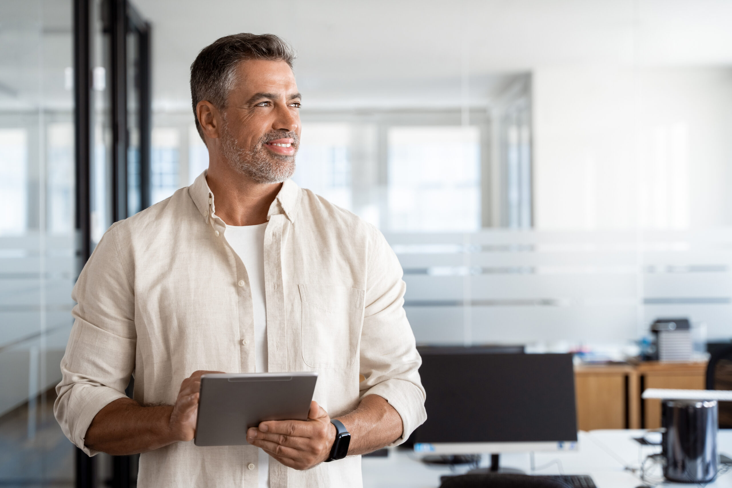 Happy Latin Hispanic bearded stylish mature adult professional business man, smiling Indian senior businessman CEO holding digital tablet using fintech tab application standing in office,looking aside